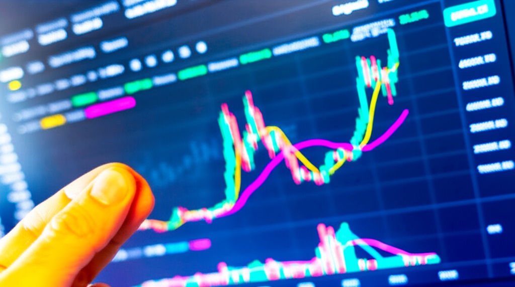 A person holding in fingers physical Bitcoin token in front of a screen showing BTC chart
